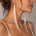 May include: A pair of long, dangling earrings featuring multiple thin, white, straight strands attached to a silver-toned metal cap. The earrings hang from a simple hook wire, extending downwards in a sleek, linear design that reaches the collarbone.